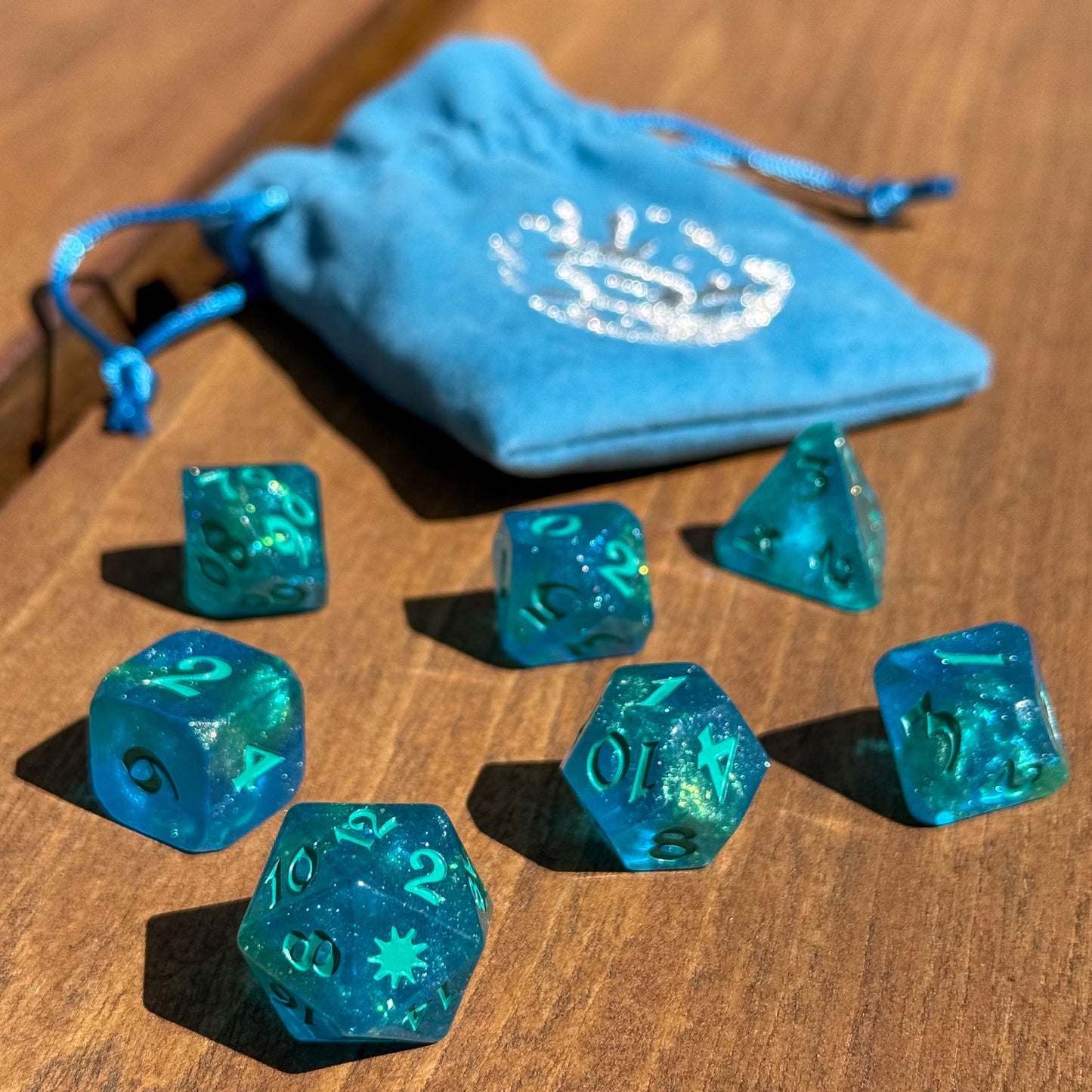 Aqua blue dice in foreground sitting on a wooden surface in natural sunlight. Velvet blue bag with silver embroidery is out of focus in the background.
