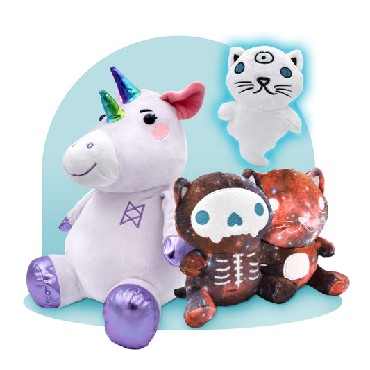 An image of four stuffed animals. On the left is a plush lavender unicorn with two rainbow horns and the Bureau of Balance logo on its chest. To the right are three plus cats, the one up top is a ghost with a third eye, the one on the far right is made of a galaxy pattern, and the one in the middle is the same galaxy pattern with a skeleton printed on top of it. 