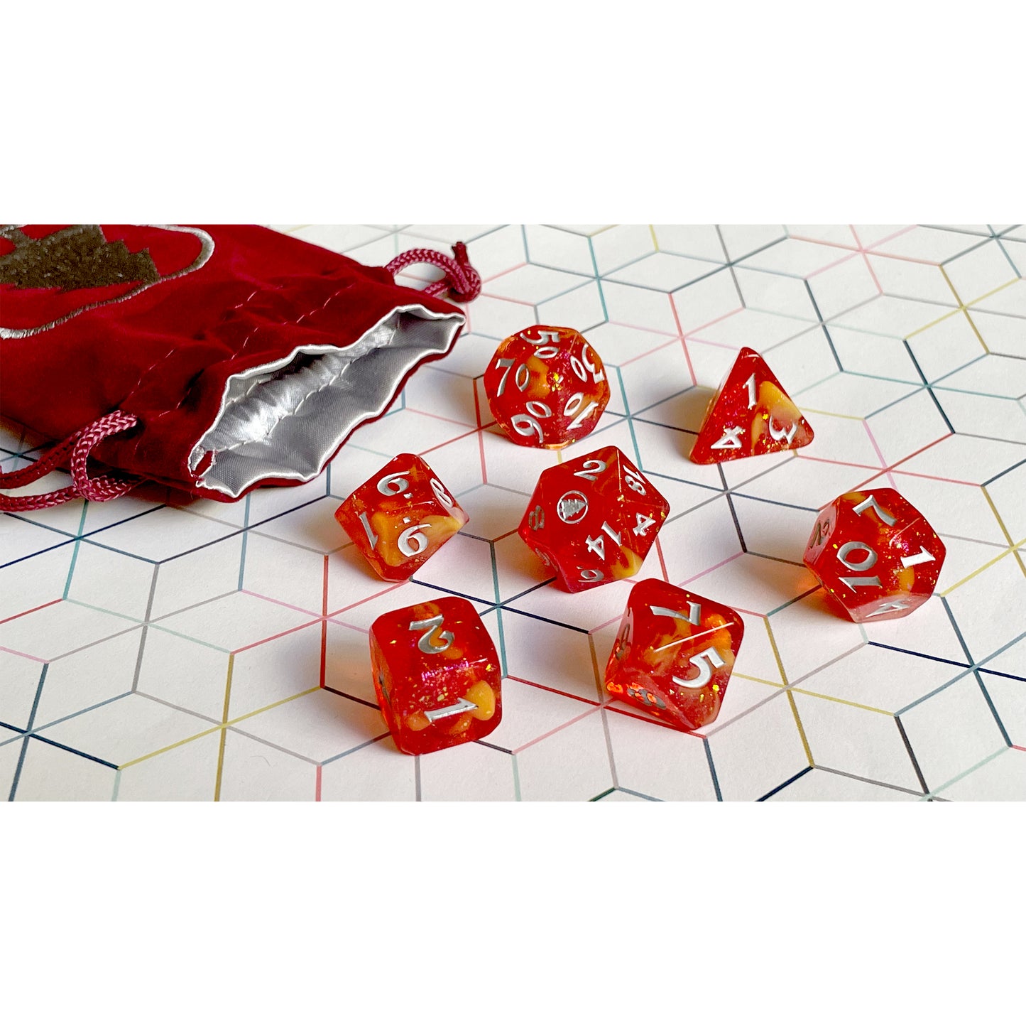 Image of an orange set of dice with silver lettering on geometric backdrop. The dice has yellow veins and glitter running through the center. The 20 on the D20 is a silver pine tree in a circle. To the left of the dice is a red velvet bag with a silver lining and the same silver pine tree emblem.