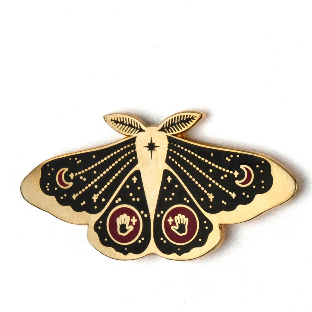 A black and gold enamel pin of a moth. Just below it's antennae is an eight pointed star. Symmetrically on both upper wings is a red crescent moon. Symmetrically on both bottom wings is a red circle with a black hand and gold cross.