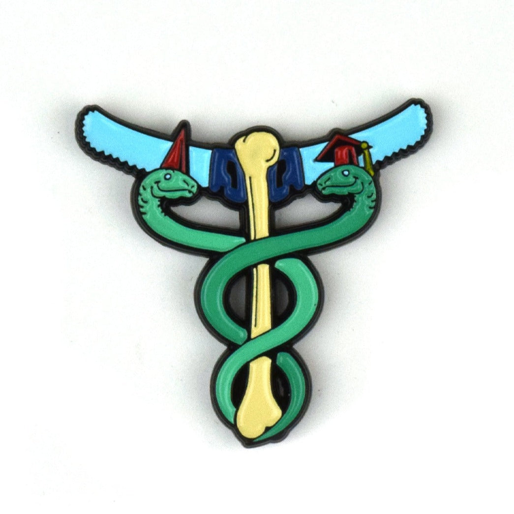 An enamel pin of two green snakes curled around a tibia bone. There is a blue saw coming out either side of the top of the bone. The snake on the left wears a conical cap and the snake on the right wears a graduation cap.