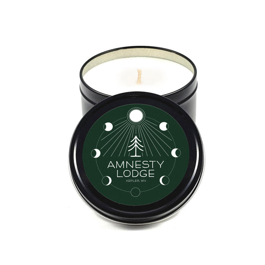A woodsy candle in a black tin. The lid has a green label with the image of a pine tree surrounded by the phases of the moon.