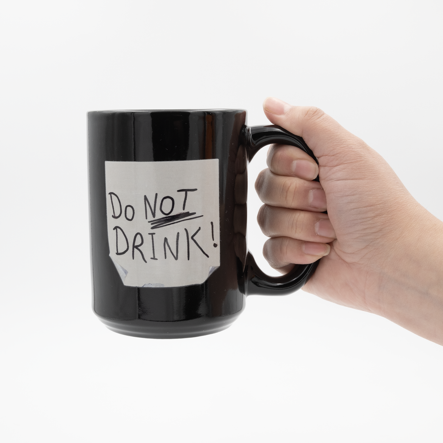 15 oz black mug with a print to look like a distressed post it note with the text DO NOT DRINK scribbled on it in black marker with its handle being held by a hand.