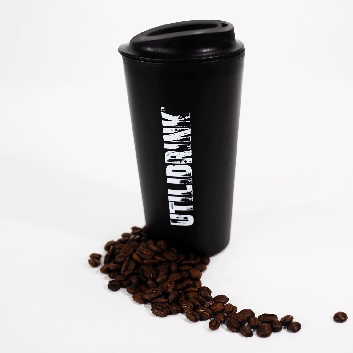 Photo of a black drink tumbler with a plastic lid. On the side it says, “UTILIDRINK” in white distressed lettering. There is a pile of coffee beans beneath it.