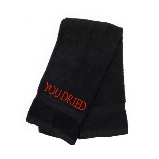 A black terrycloth hand towel on a white background, folded twice. At one end of the towel is bright red all caps letters reading “You dried”.