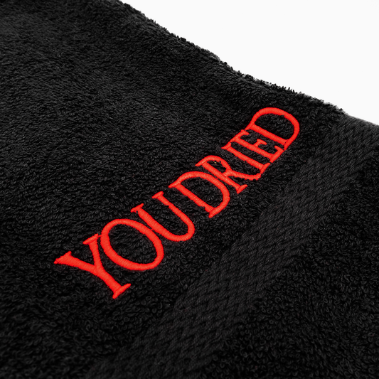 A close up photo of black terrycloth with a braided black band. Just above the band is bright red embroidered all caps text that reads "You dried".