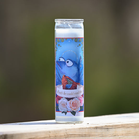 Photograph of a tall white candle in a glass container with a sticker applied to the front. The sticker shows a cartoon blue fish with a halo holding up one fin in a wave, wearing a red draped fabric and holding a book that looks like it might say "learn to read". A banner across the middle says "if only he could read this" and below the banner are four pink and red roses.