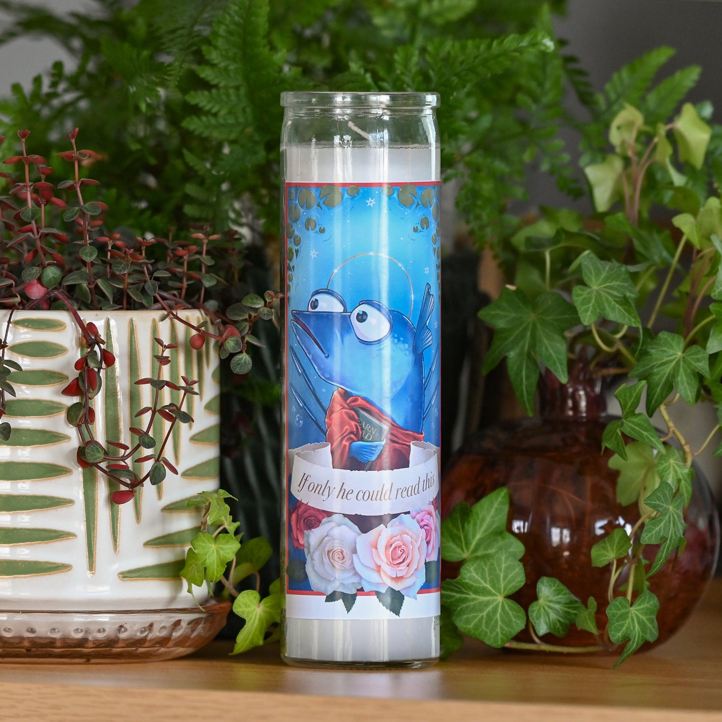 Photograph of a tall white candle in a glass container with a sticker applied to the front. The sticker shows a cartoon blue fish with a halo holding up one fin in a wave, wearing a red draped fabric and holding a book that looks like it might say "learn to read". A banner across the middle says "if only he could read this" and below the banner are four pink and red roses. 

Behind the candle and out of focus are various potted plants and vases.