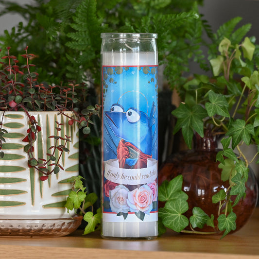 Photograph of a tall white candle in a glass container with a sticker applied to the front. The sticker shows a cartoon blue fish with a halo holding up one fin in a wave, wearing a red draped fabric and holding a book that looks like it might say "learn to read". A banner across the middle says "if only he could read this" and below the banner are four pink and red roses. 

Behind the candle and out of focus are various potted plants and vases.