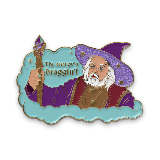 Pin with gold plating of white haired man (Clint) dressed in a purple wizards robe holding a wooden staff with crystals. The underside of his hat and one of the crystals has purple glitter enamel. The background is a blue cloud with gold text that says, "The Energy's Draggin'!"