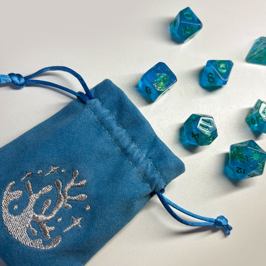 Light blue velvet dice bag with silver embroidery of a coral laid on flat white surface. Blue dice with green glitter lay on the flat surface in front of the bag.