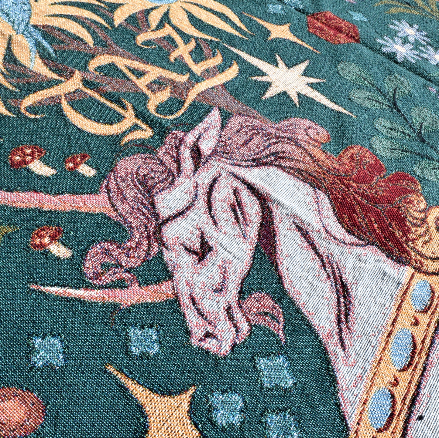 closeup of a deep green woven blanket with gold stars, red and white mushrooms, and flowers on it. in the center, a unicorn's head with two long reddish horns.