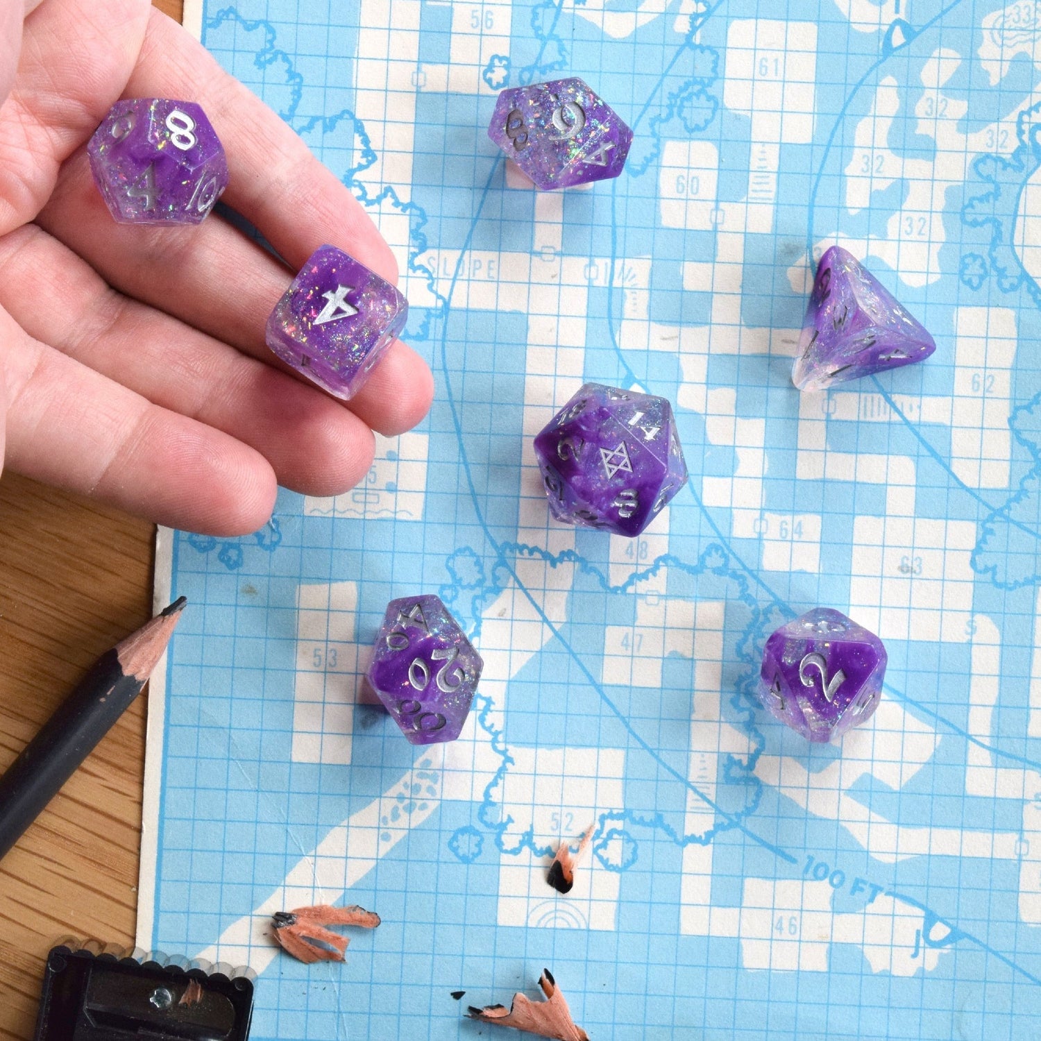 Image of a seven dice set: a D4, D6, D8, D10, D12, D20, and D100. The dice have a galaxy swirl inside them of dark purple, light purple, and clear with iridescent glitter flakes. The numbers are painted silver. The D20 has the BoB logo on the 20 side. This scene shows them being rolled on a graph paper RPG map.