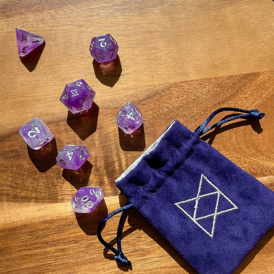 Image of a seven dice set: a D4, D6, D8, D10, D12, D20, and D100. The dice have a galaxy swirl inside them of dark purple, light purple, and clear with iridescent glitter flakes. The numbers are painted silver. The D20 has the BoB logo on the 20 side. Also shown is a deep purple velvet dice bag with draw string. It has silver embroidery in the shape of the BoB symbol. 
