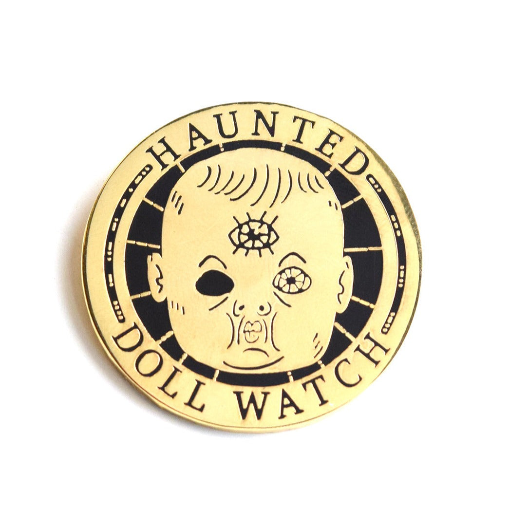 A gold and black enamel pin. In the center is a doll's head. It's missing it's left eye but has a third eye on its forehead. Around the outside it says, "Haunted doll watch".