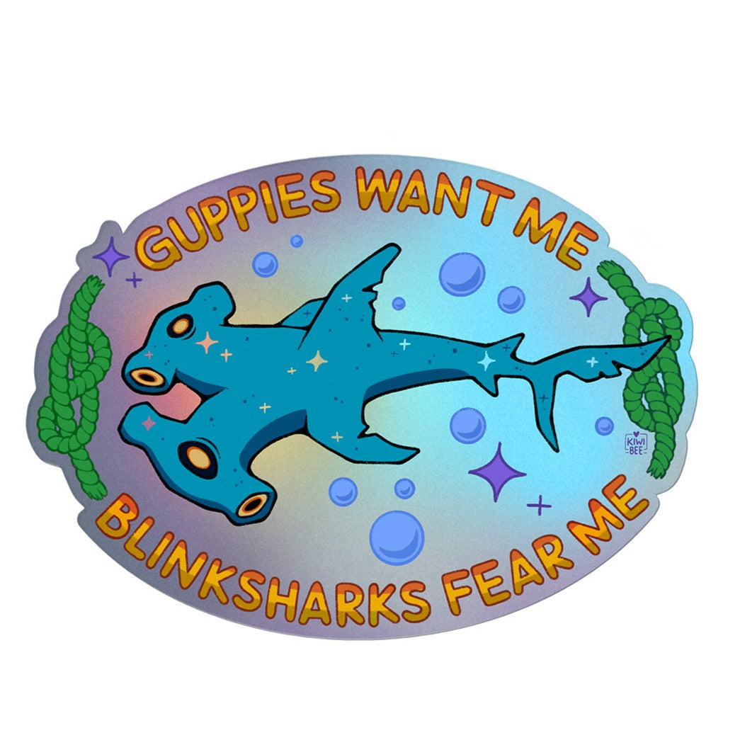 An oval shaped sticker with an iridescent background. At the top is says, “Guppies want me” and at the bottom it says, “Blinksharks fear me”. In the center is a blue two-headed shark surrounded by bubbles and sparkles. On either side of the shark is a green sailor’s knot. 