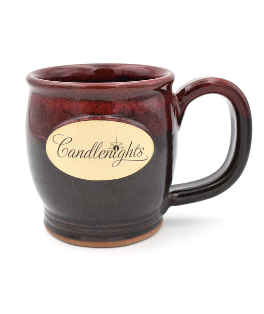 A round stoneware mug. The mug fades from a maroon to dark brown glaze. There's a cream oval in the center that says, "Candlenights" in script. The "i" is a lit candle.