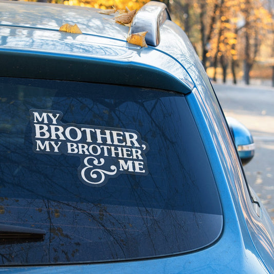 A photograph of the back window of a blue car. There are some yellow leaves on the roof of the car. There is a decal on the back window that says, "My Borther, My Brother & Me" is a white serif font.