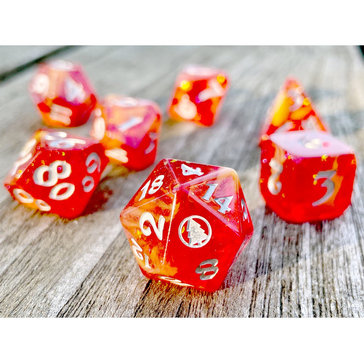 Image of an orange dice set with silver lettering. The dice have yellow veins and glitter running through the center of them. The 20 on the D20 is a silver pine tree in a circle. 