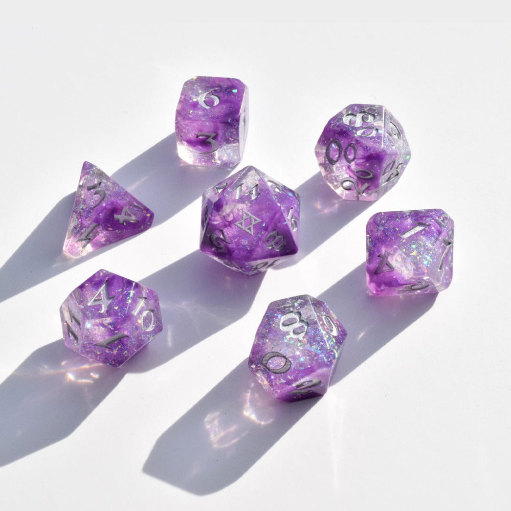 Image of a seven dice set: a D4, D6, D8, D10, D12, D20, and D100. The dice have a galaxy swirl inside them of dark purple, light purple, and clear with iridescent glitter flakes. The numbers are painted silver. The D20 has the BoB logo on the 20 side. 