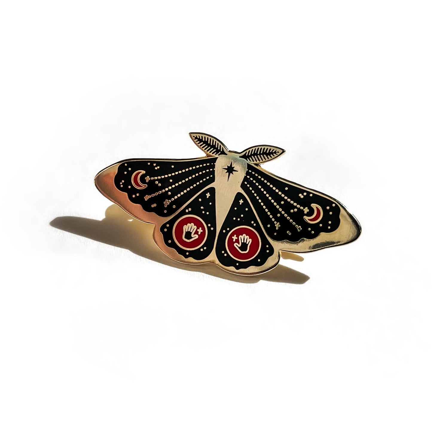 A black and gold enamel pin of a moth. Just below it's antennae is an eight pointed star. Symmetrically on both upper wings is a red crescent moon. Symmetrically on both bottom wings is a red circle with a black hand and gold cross.