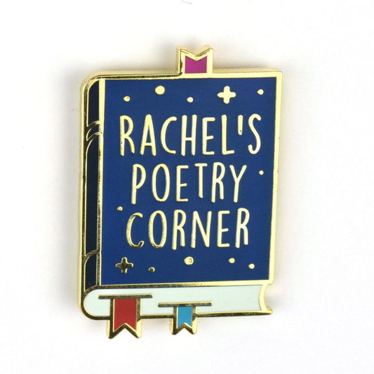 A gold enamel pin of a dark blue book with three bookmarks: one purple, one red, and one blue. On the cover it says "Rachel's poetry corner" in gold with gold dots and crosses.