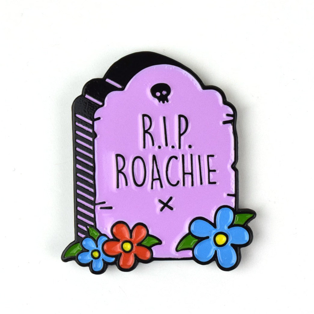 An enamel pin of a light purple grave stone with red and blue flowers. It says, "R.I.P. Roachie X" with a small black skull above it. 