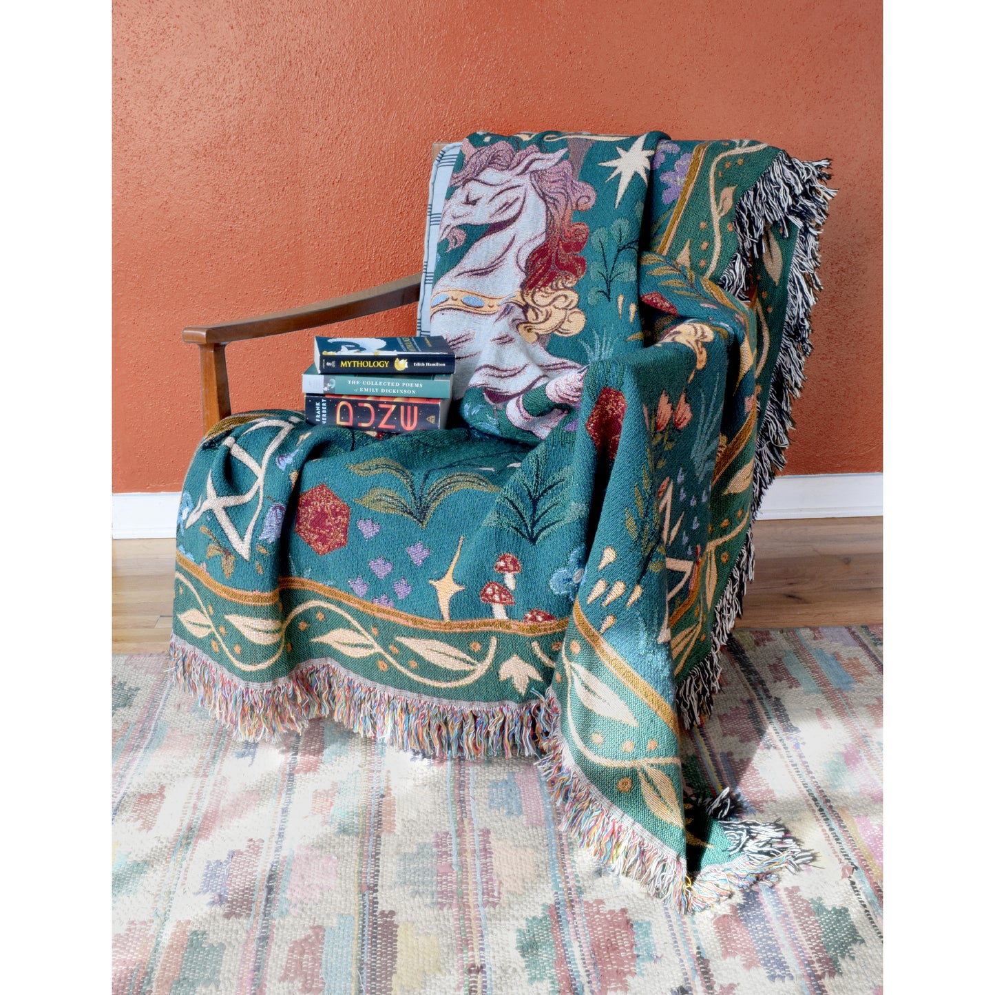Photo of a teal and green tapestry blanket with Garyl in the center. He’s rearing back on his hind legs and has a flowing mane and tail. There are flowers around him. Above him is a tree with blue birds. There is a B.o.B. rune in each corner. The blanket is draped over a wooden chair with a stack of books on it. Sunlight streams into the room, highlighting a soft tan patterned rug and terracotta coloured wall. 