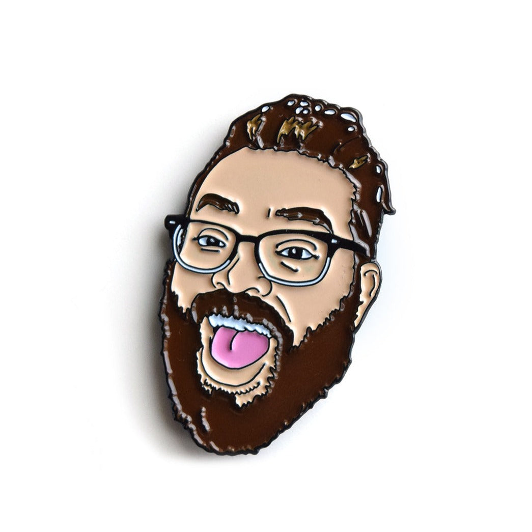 An enamel pin of Travis's head with his mouth open. He has brown hair, a thick beard, and black and white glasses.